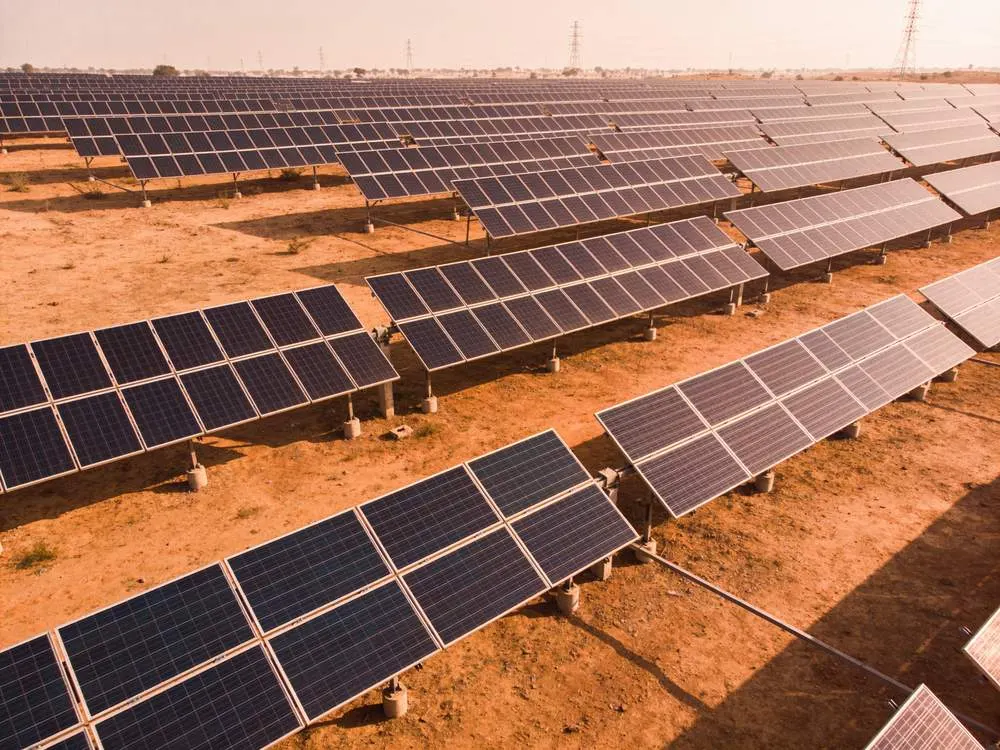 Weite Reihen von Solarpaneelen auf rötlichem Wüstenboden, im Hintergrund Strommasten unter blassem Himmel.